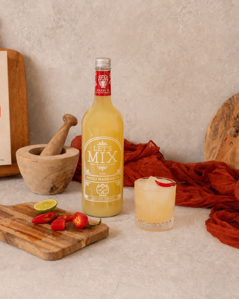 Salt-rimmed Chilli Margarita with fresh chillies and lime next to a bottle of Let's Mix Chilli Margarita Mix